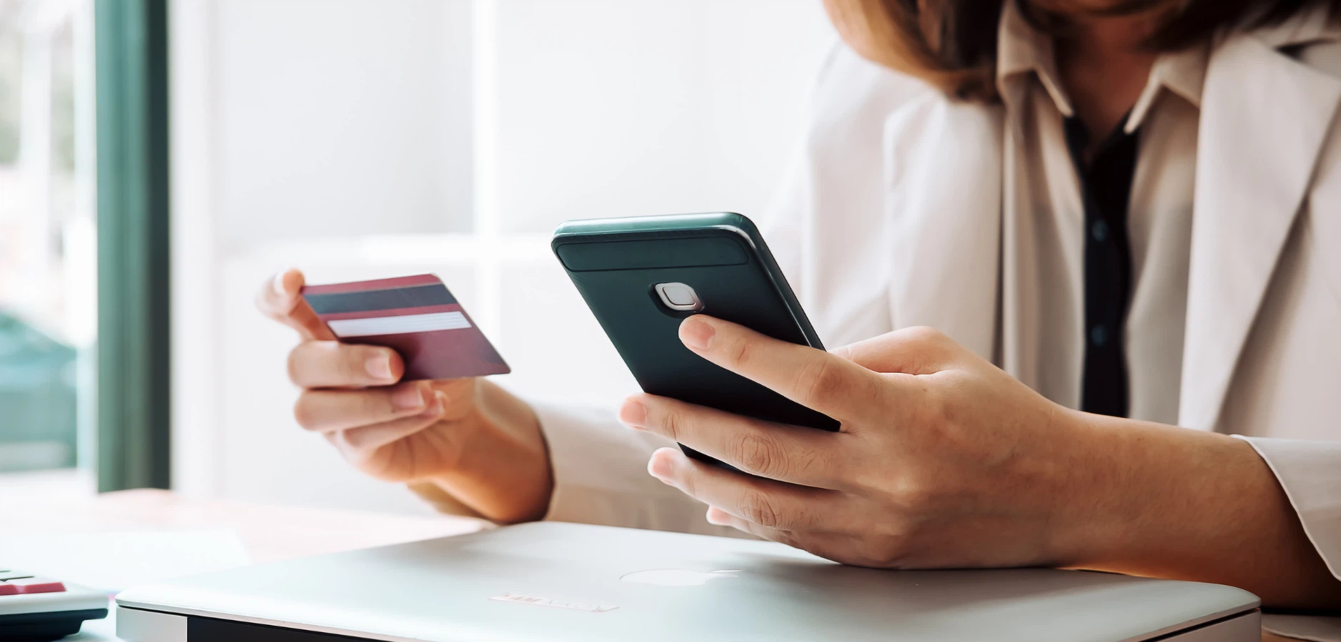 A person in a white blazer holds a smartphone in one hand and a credit card in the other, sitting at a desk with a laptop.