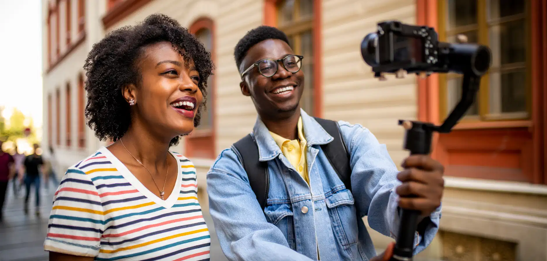 A young man holds a camera on a stabilizer, capturing a moment with a woman in colorful attire outdoors, surrounded by urban scenery.
