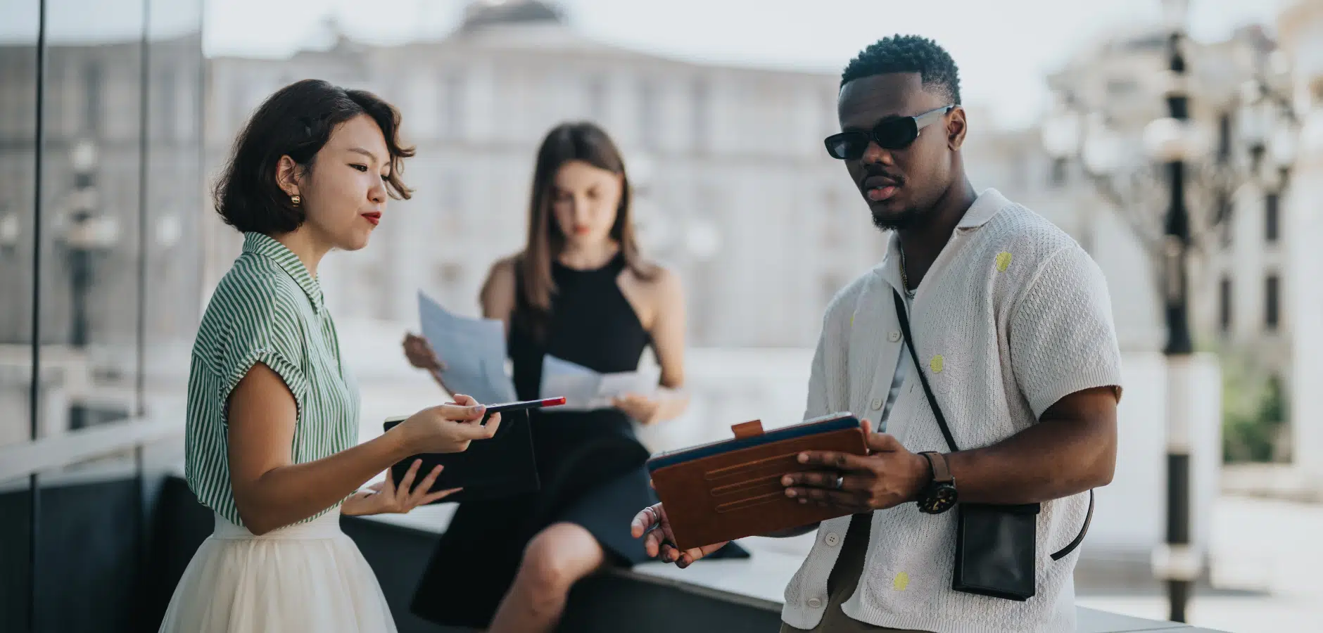 A group of three people engaged in discussion outdoors, holding documents and a tablet, against an urban backdrop.