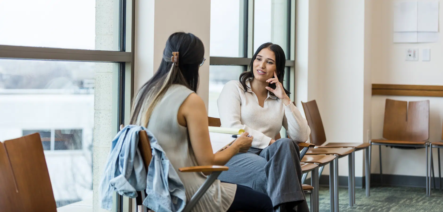 Two women are seated in a bright waiting area, engaged in conversation, with wooden chairs and large windows in the background.