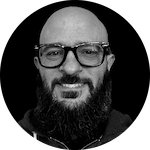 A person with a full beard and a bald head wearing a dark hoodie, set against a solid black background.